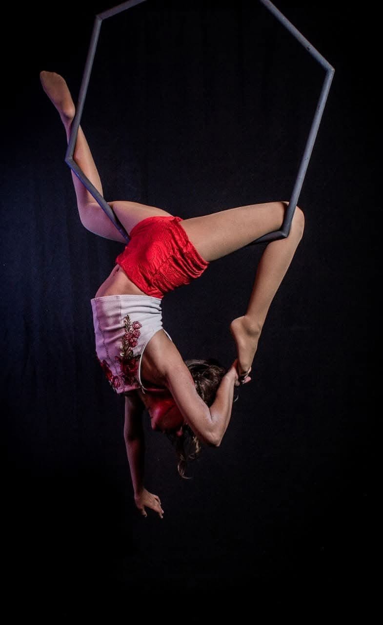 Florencia invertida en un hexágono de metal, con una pierna extendida, sobre fondo negro