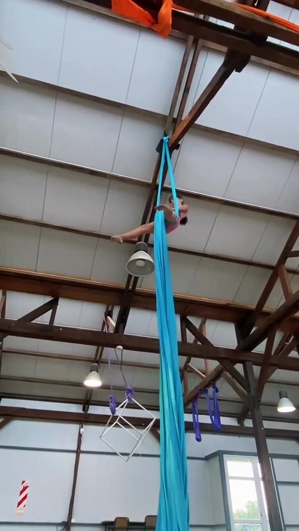 Alumna en una tela aérea turquesa, a varios metros de altura, bajo el techo de vigas de una nave