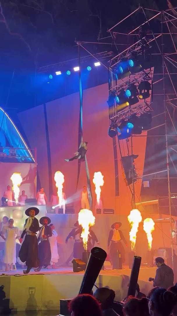Acróbata en una tela aérea sobre un escenario, con llamaradas y bailarines en escena